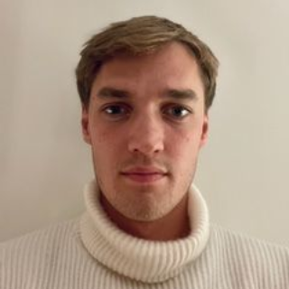 Bendik Grieg Skjold med lys hud og brunt hår iført en hvit polotrendgenser, mot en hvit vegg.