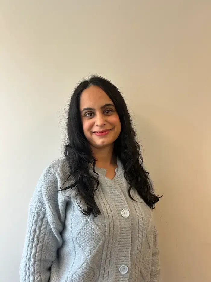 Kvinne med mørkt hår, iført en grå kabelstrikket cardigan, smiler mot en nøytral vegg.