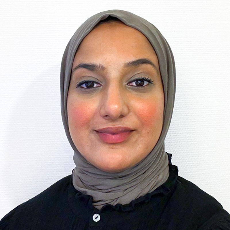 Woman with brown hijab and black top smiles, lit by natural light.