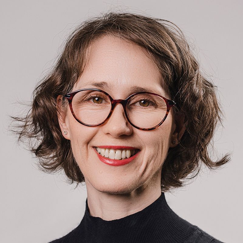 Woman with glasses, smiling, wearing a black top, against a gray background.