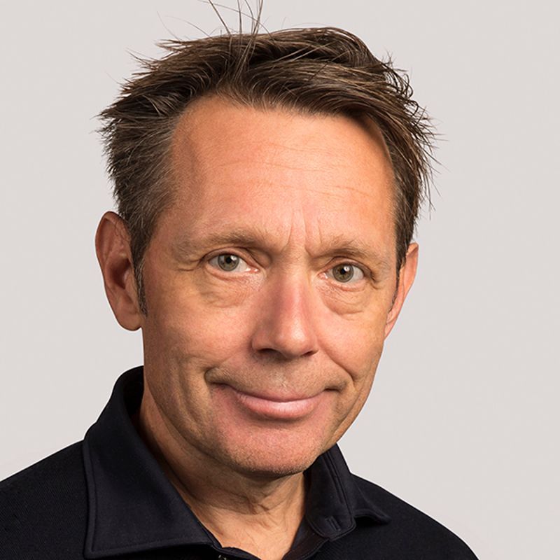 Middle-aged man with light brown hair, smiling slightly, wearing a dark blue shirt, against a neutral background.