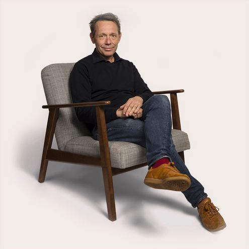 Man in black shirt and jeans sitting in a gray armchair, brown shoes, one leg crossed, looking towards the camera.