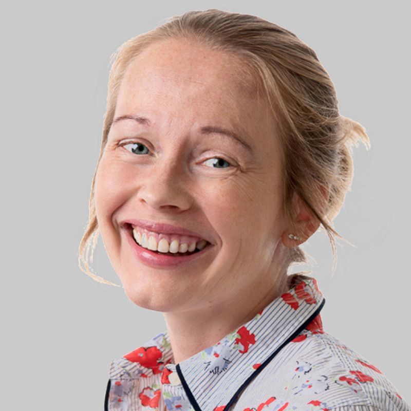 Smiling blonde woman wearing a floral shirt with a light gray background.