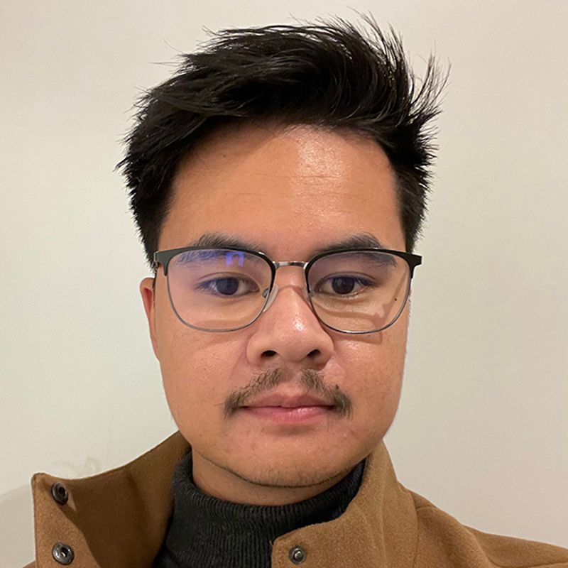 Man with glasses, dark hair, and a mustache, wearing a tan jacket and gray turtleneck, looking at the camera.