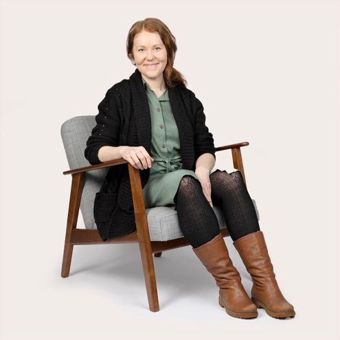 Woman with red hair, smiles, sits in a gray chair, wearing black cardigan, green dress, and brown boots.