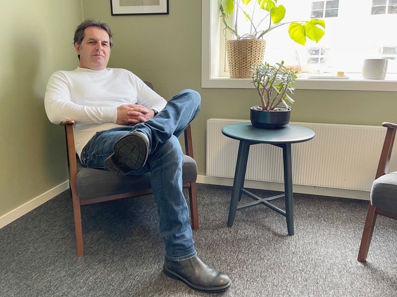 Man sitting in a chair, legs crossed, in a room with a small table and plants.