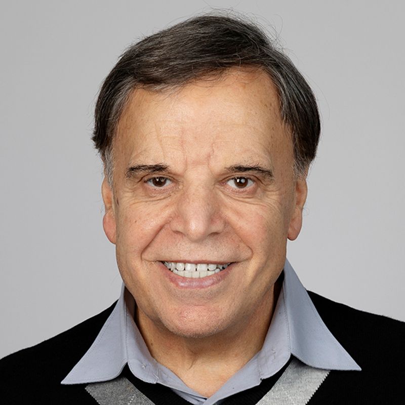 Smiling older man with dark hair, wearing a light blue collared shirt under a black sweater.