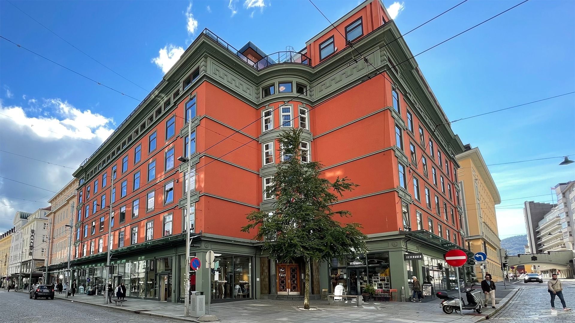 Red multi-story building with shops on ground floor. Corner view, sunny day, pedestrians on street.
