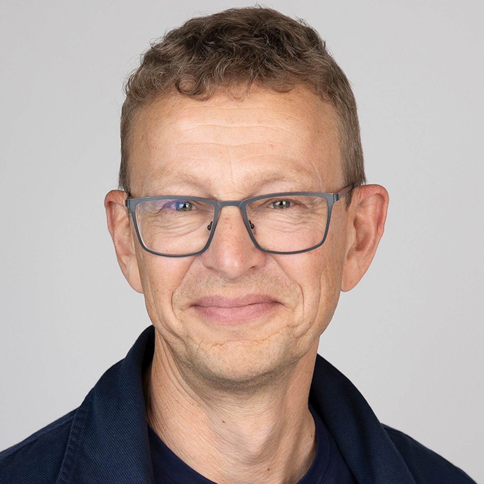 Man with glasses smiling, wearing a blue jacket, against a white background.