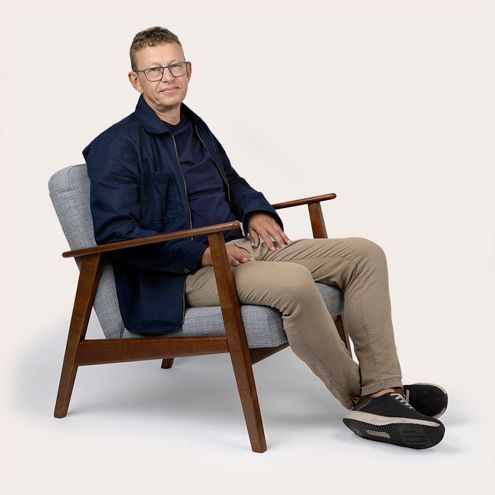 Man in glasses sits in a grey chair; wearing a navy jacket, tan pants, and black shoes.