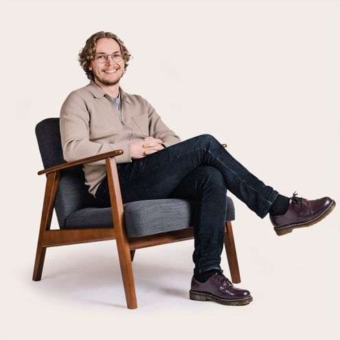 Man with curly hair, smiles while sitting in a mid-century chair, wearing a beige sweater, jeans and purple shoes.