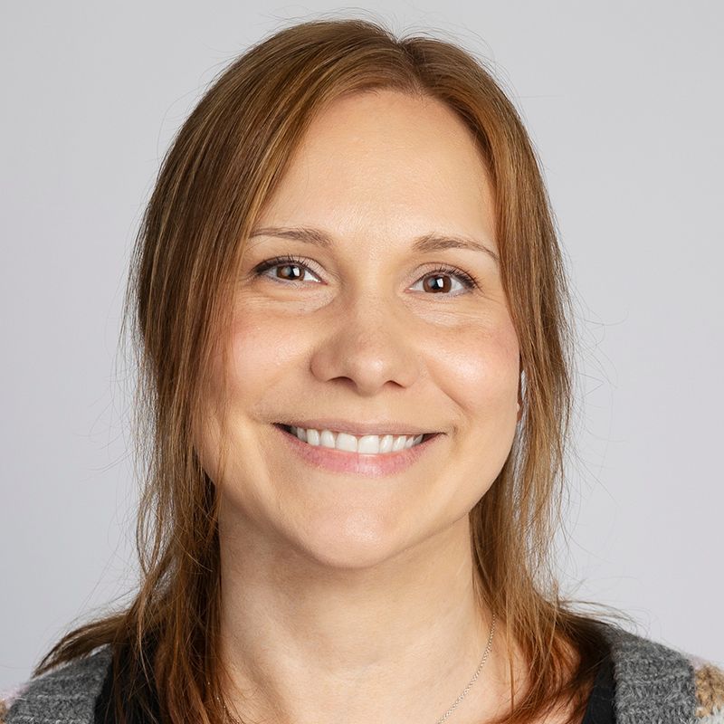 Woman with light brown hair, smiling, wearing a gray and patterned sweater, against a gray background.