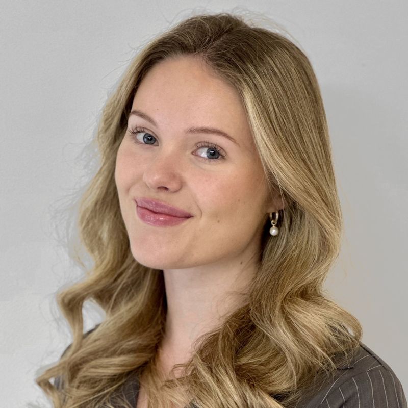 Blonde woman with wavy hair smiles at the camera. She wears a blazer and pearl earrings.