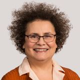 Woman with curly brown hair and glasses smiles, wearing an orange top and cream shirt.
