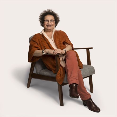 Woman with curly hair smiles while seated, wearing rust-colored outfit and brown boots.