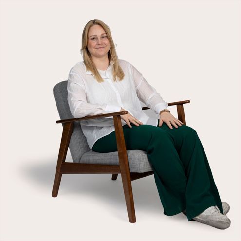 Woman with blonde hair, wearing a white shirt and green pants, seated in a gray chair, smiling.