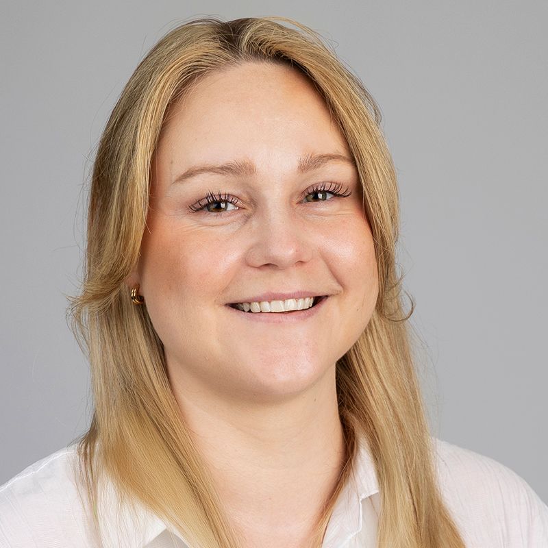 Blonde woman smiling at the camera, wearing a white shirt against a gray background.