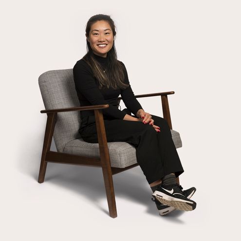 Woman with dark hair smiles, seated in a gray chair, wearing black clothing and sneakers.