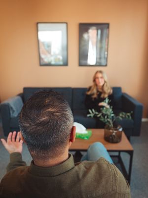 Man facing away gestures toward woman on sofa in therapy session.