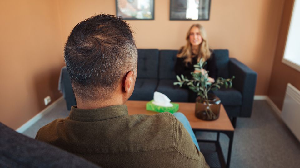 Man in foreground facing a smiling woman on a couch. Therapist's office setting with tissue box.