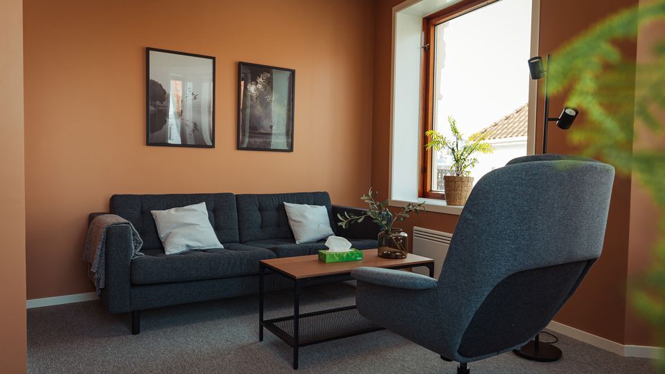 A cozy room with a dark gray couch, a matching chair, and a small table; framed pictures on the orange wall.