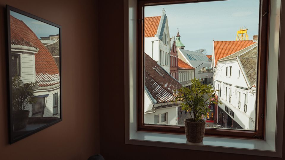 Window view of a narrow street with red-tiled roofs and white buildings, a potted plant on the windowsill, and a framed picture.