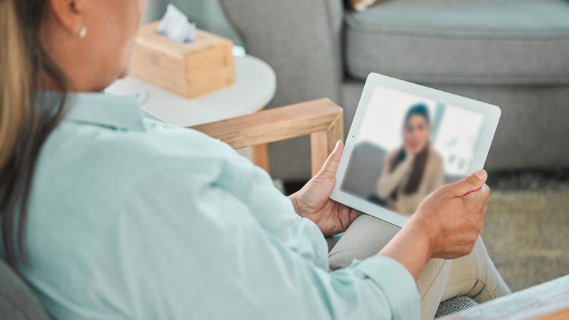Woman in mint shirt on floor, using tablet for video call with therapist.