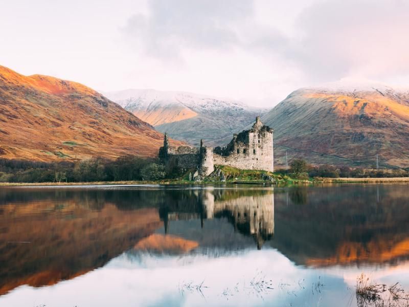 Ruined castle on a lake reflects the surrounding golden hills and overcast sky.