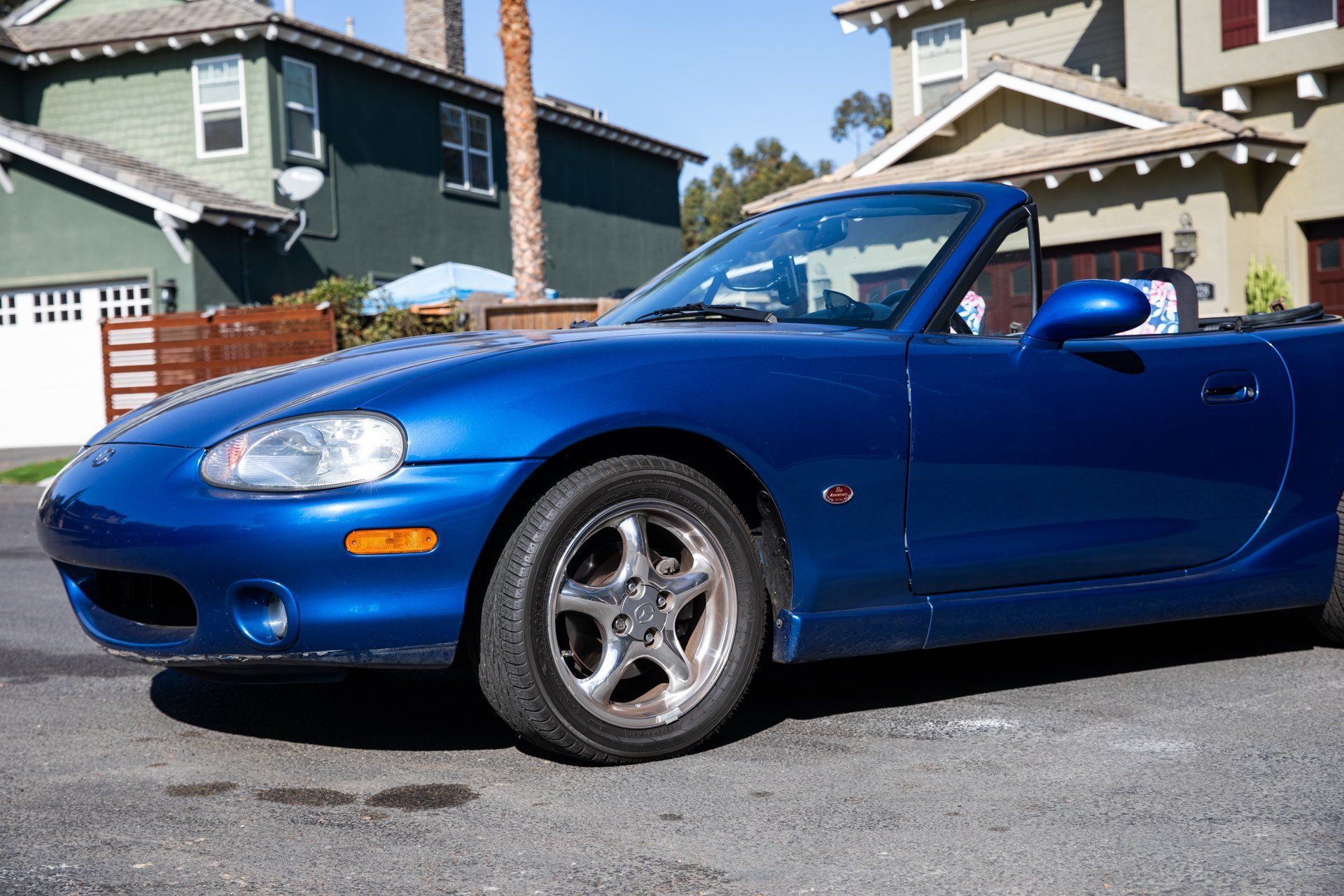 A blue Mazda Miata convertible parked on a suburban street in front of two-story houses.