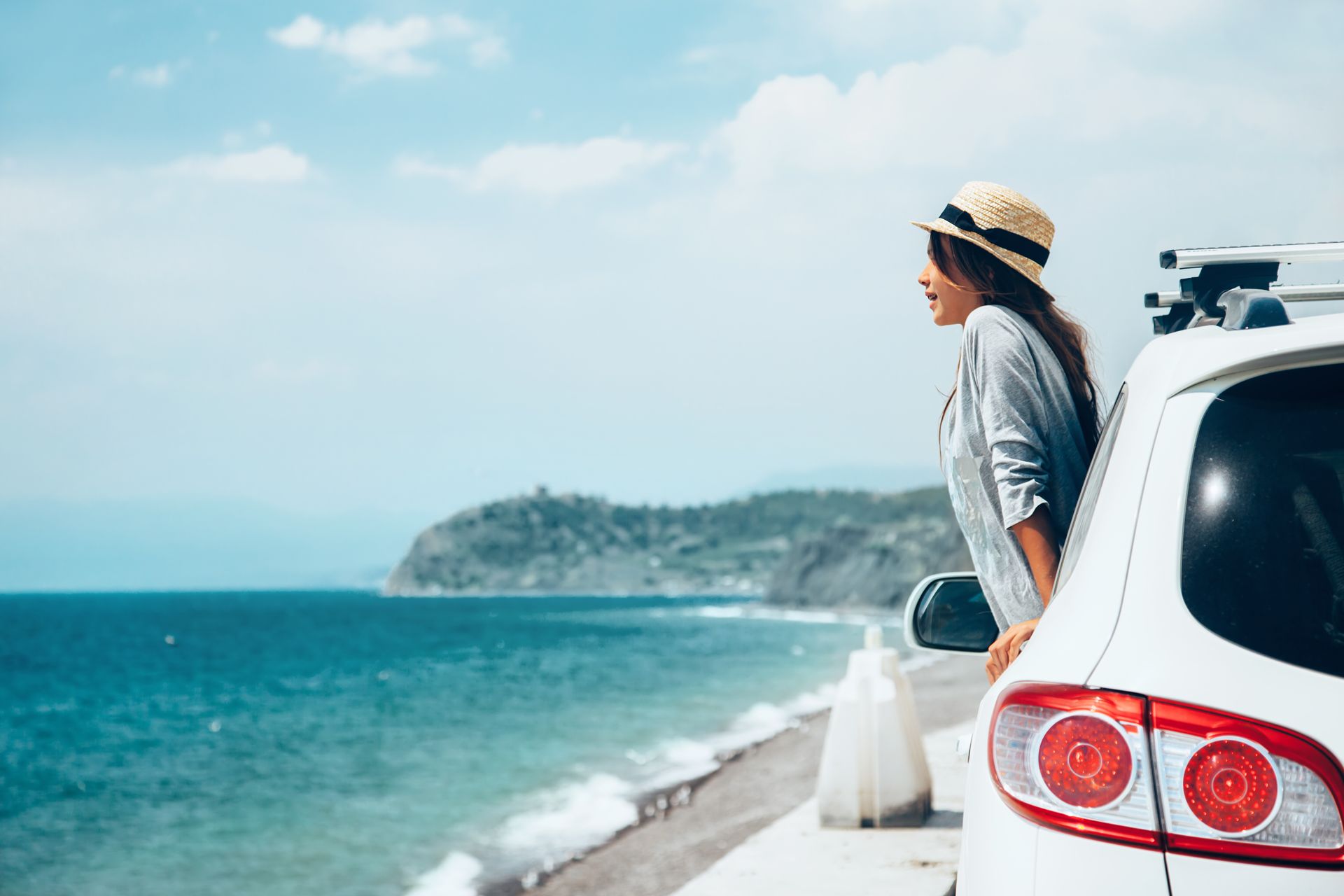 Woman in straw hat looks out at ocean from a white car parked on a coastal road.