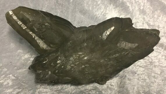 A Dark, Smokey Quartz Crystal With a Rough, Jagged Surface Lies on a Gray Textured Background — Crystal Cauldron In South Mackay, QLD