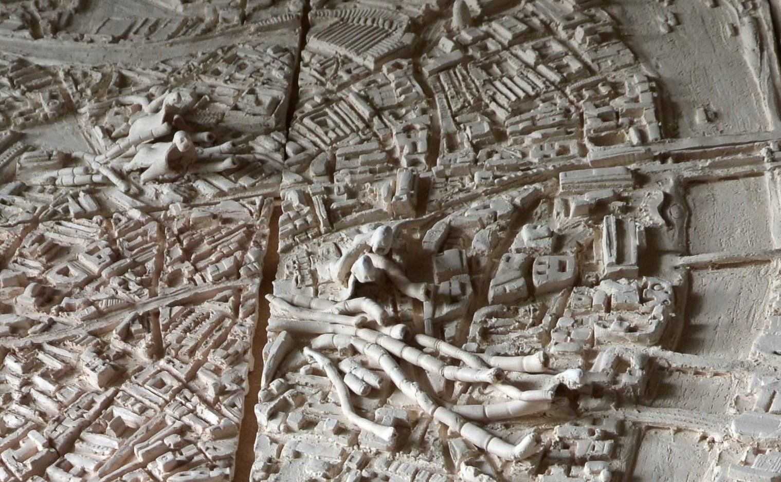 Een beige, gestructureerde reliëfkaart van een stadsplattegrond met gebouwen, wegen en waterwegen, verdeeld in panelen.