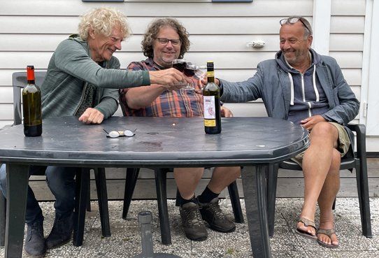 Drie mensen zitten aan een tafeltje buiten, glimlachen en klinken met hun glazen rode wijn om te proosten.
