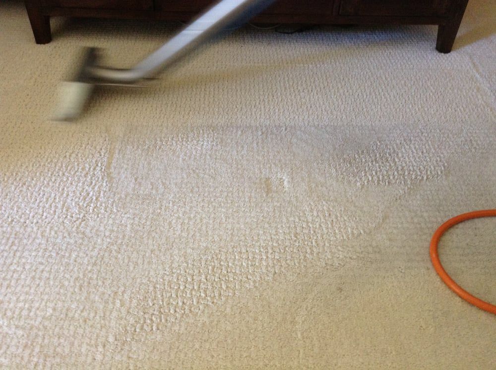 Carpet being vacuumed under a wooden table, with a beige vacuum head and orange hose visible.
