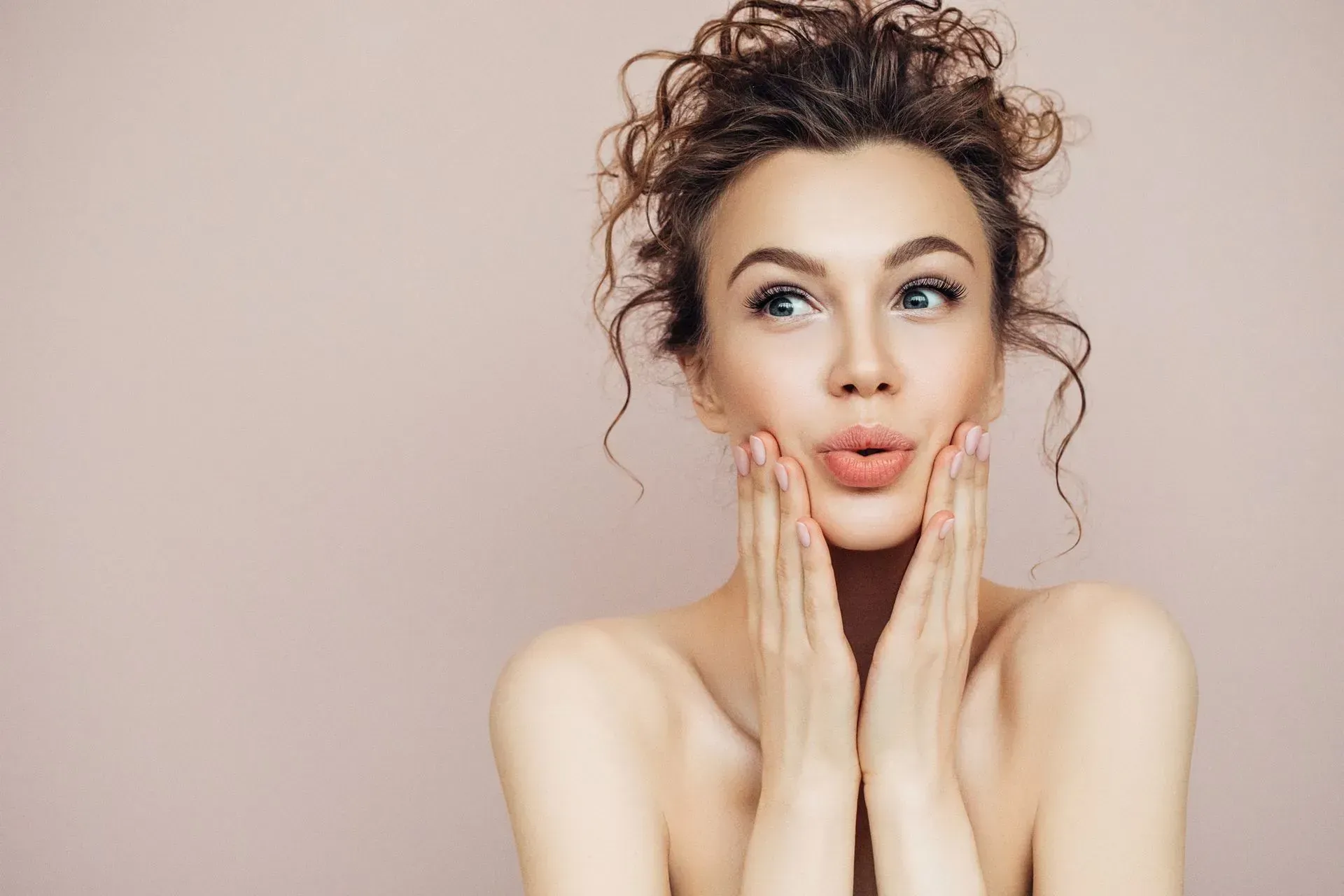 Woman with curly hair and bare shoulders touching her cheeks against a soft pink background