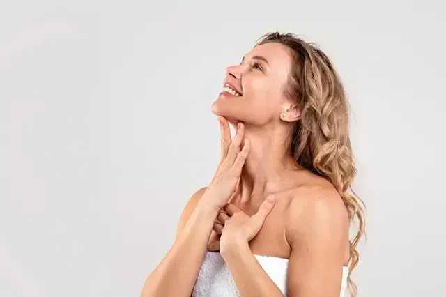 Smiling woman touching her neck and jaw, wrapped in a white towel against a light background