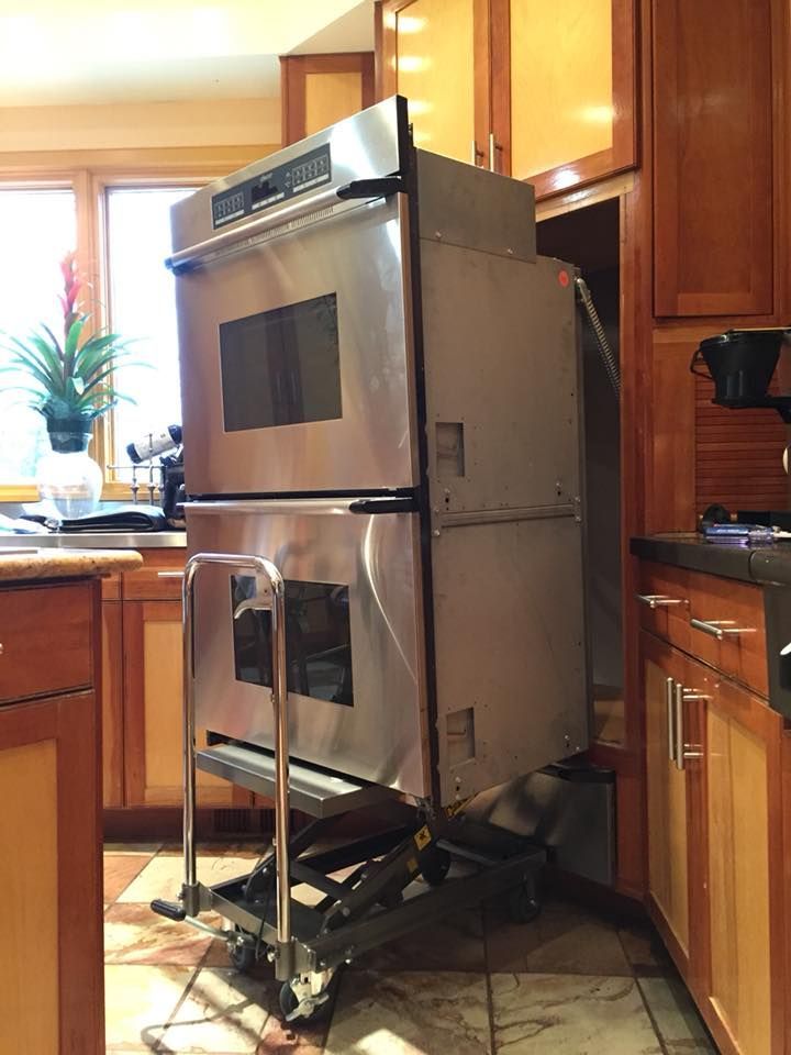 Double oven on a lift cart being installed in a kitchen with brown cabinets and a window.