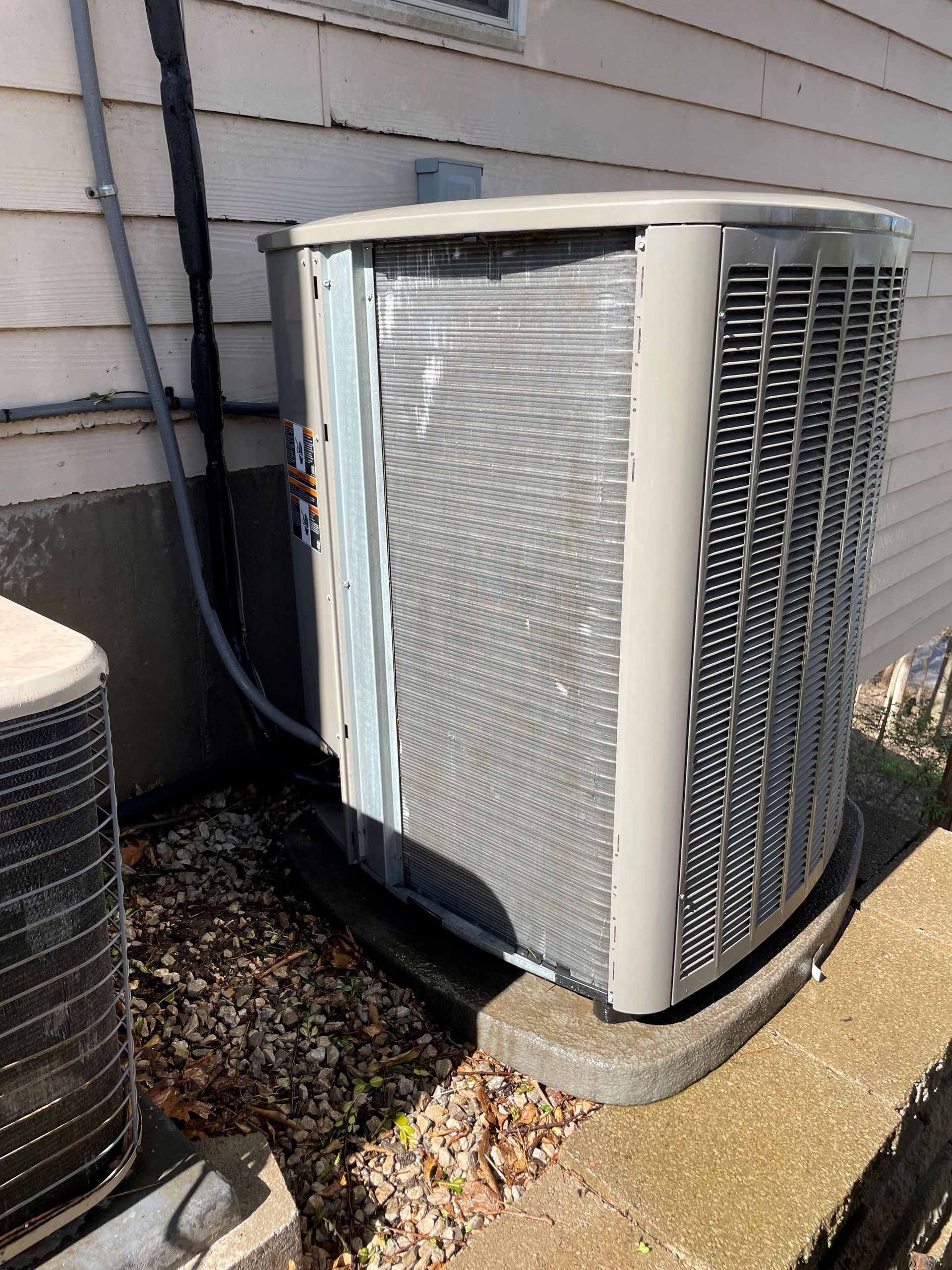 An outdoor air conditioning unit, gray and metallic, sits on a concrete pad near a house.