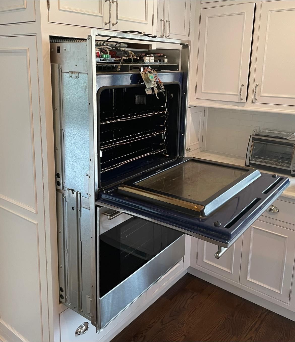 Oven removed from cabinetry, door open. Stainless steel interior. White cabinets and dark wood floor.