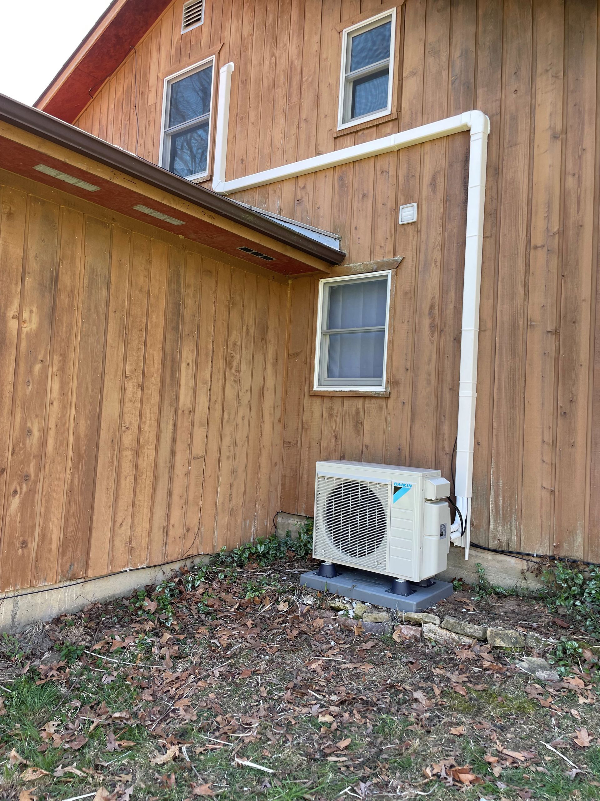 Exterior of a wooden building with an AC unit outside on a concrete pad. White piping runs up the wall.