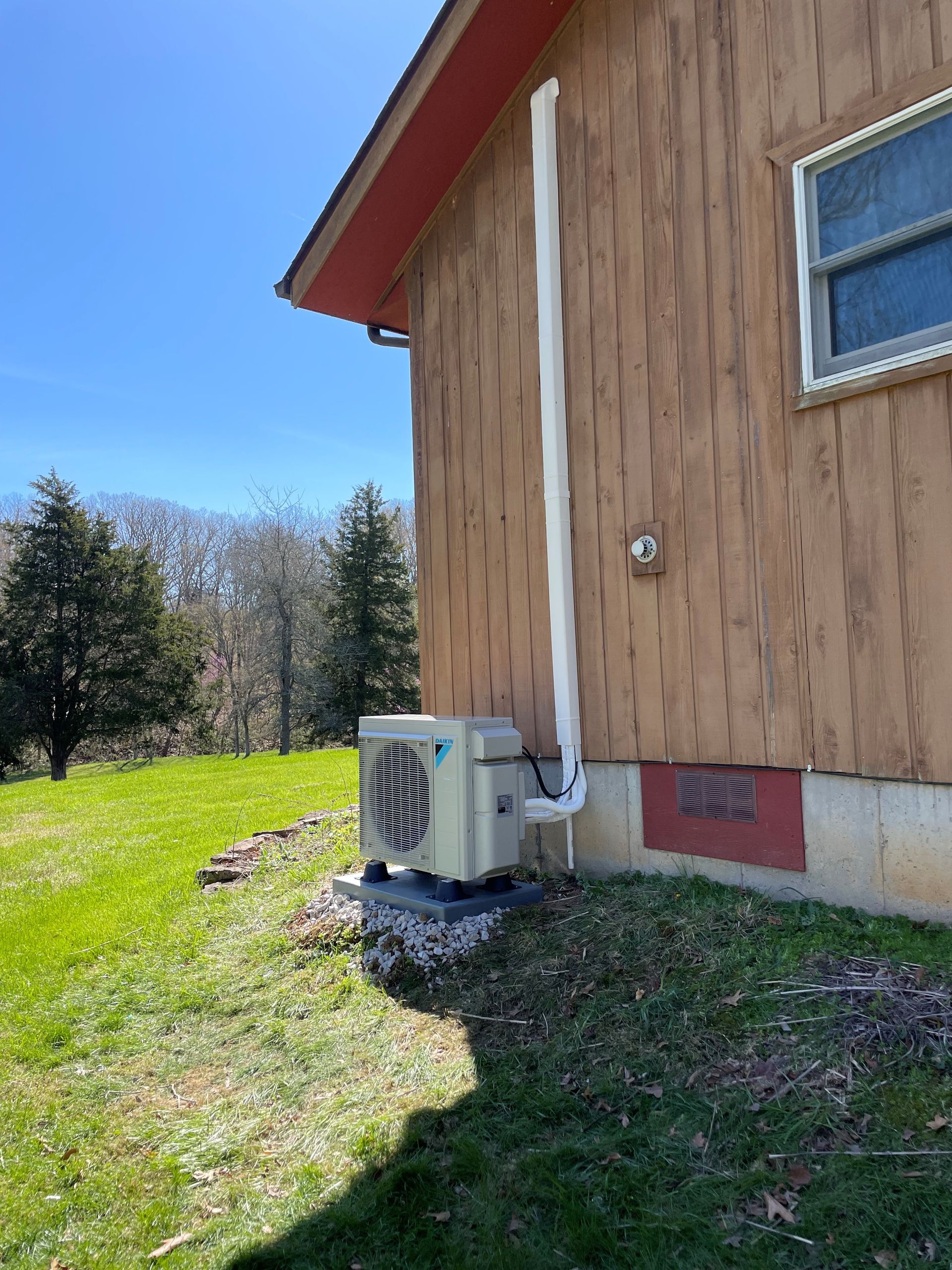 An air conditioning unit next to a wooden building with white piping against a blue sky.