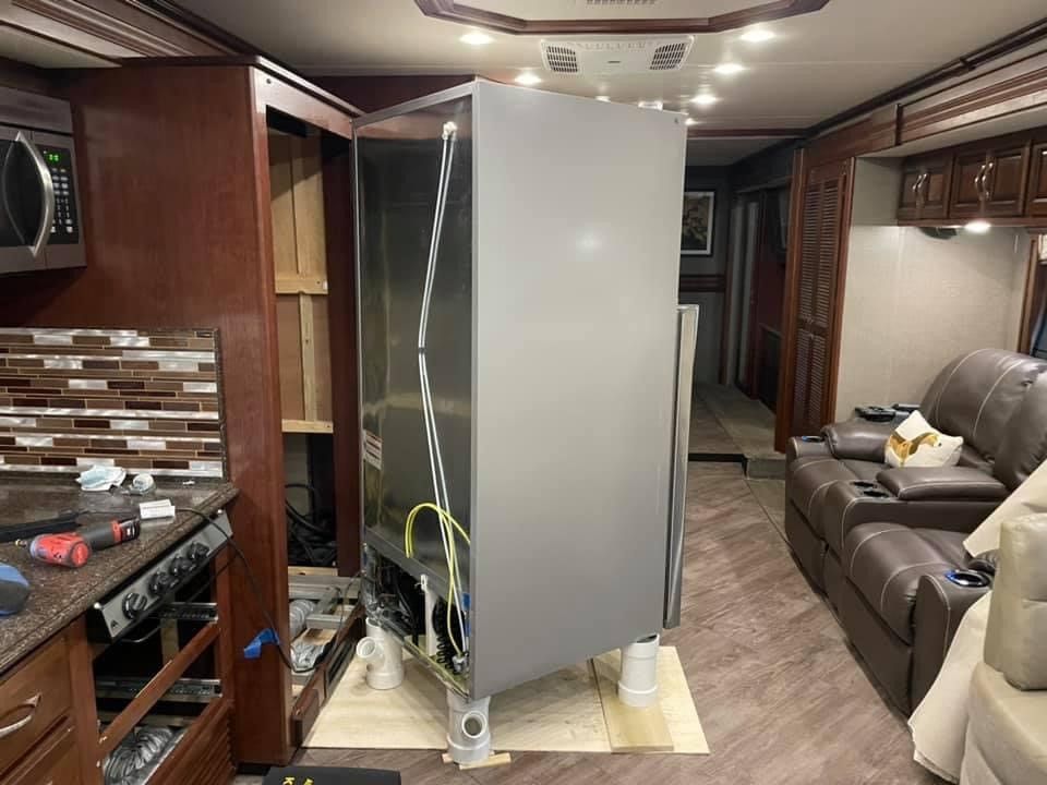 Refrigerator being removed from RV, interior view. Gray appliance, wooden cabinets, brown floors, and sofa visible.