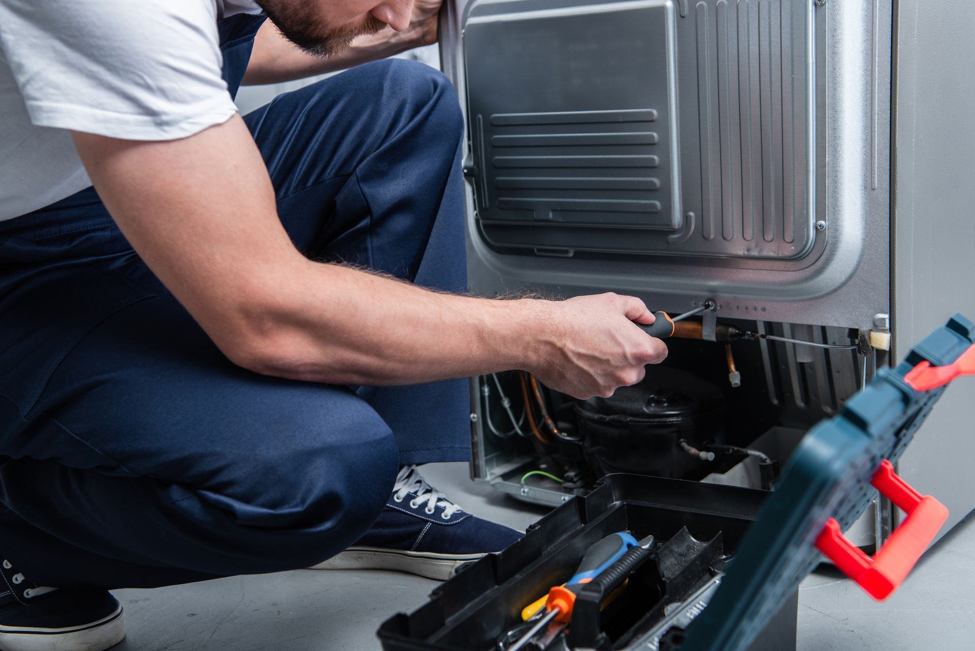 repairman in working overall fixing refrigerator by screwdriver in kitchen. repairman in working overall fixing refrigerator by screwdriver in kitchen.