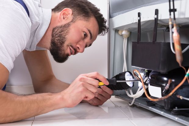 Professional technician performing fridge repair using flashlight while inspecting compressor. 