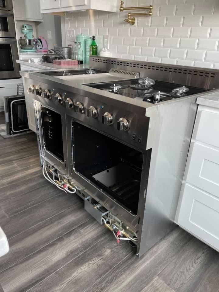 Stainless steel oven range being installed in a kitchen with gray flooring and white cabinets.