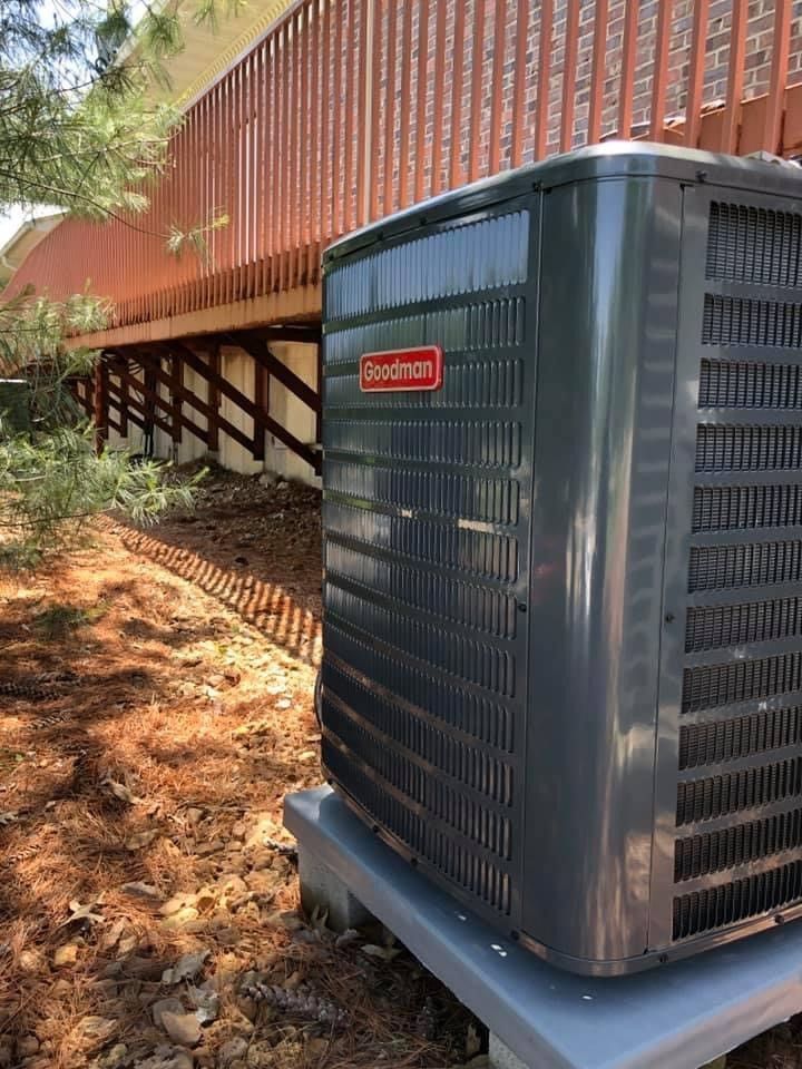 Gray Goodman air conditioner unit next to a wooden deck on a bed of pine needles.