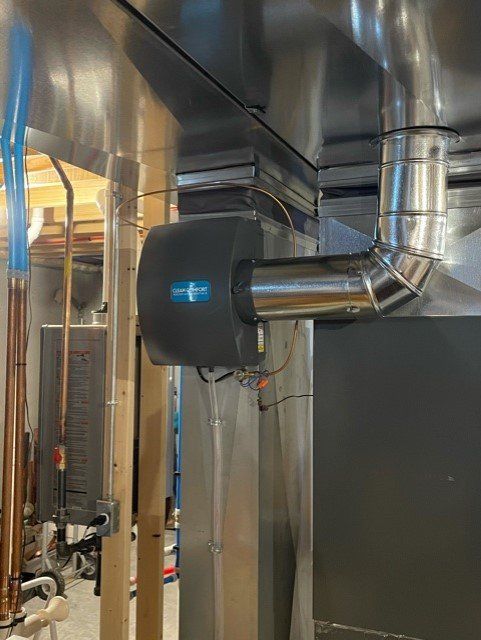 A humidifier attached to ductwork in a basement. It's a dark gray unit with a silver pipe, next to metal ducts and pipes.