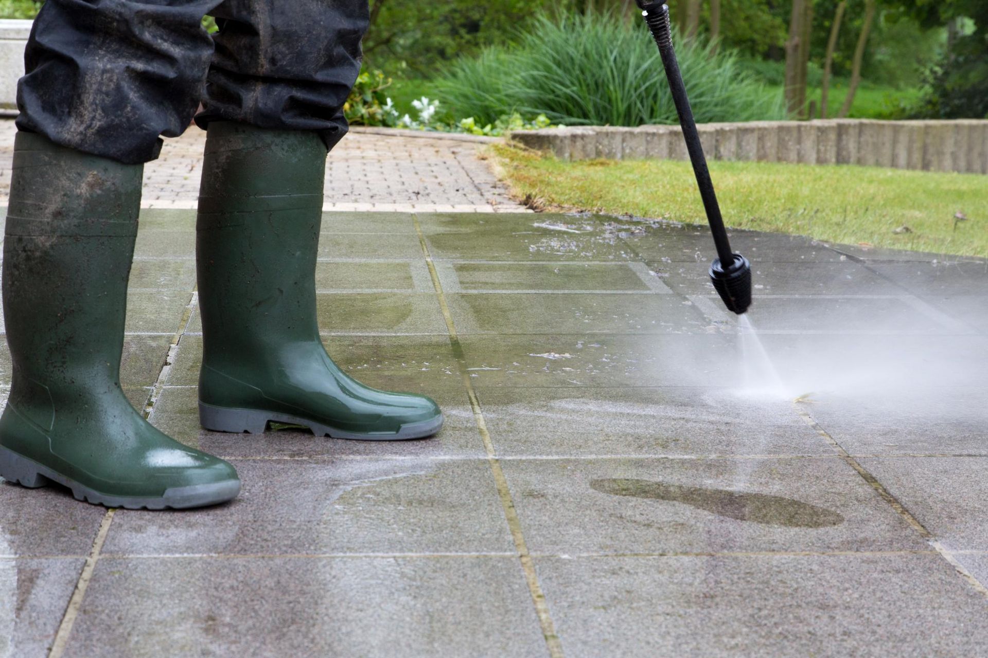 Person in green boots pressure washing a stone patio outdoors.