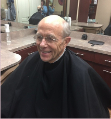 Smiling elderly man with glasses, wearing a black cape, at a barber shop.