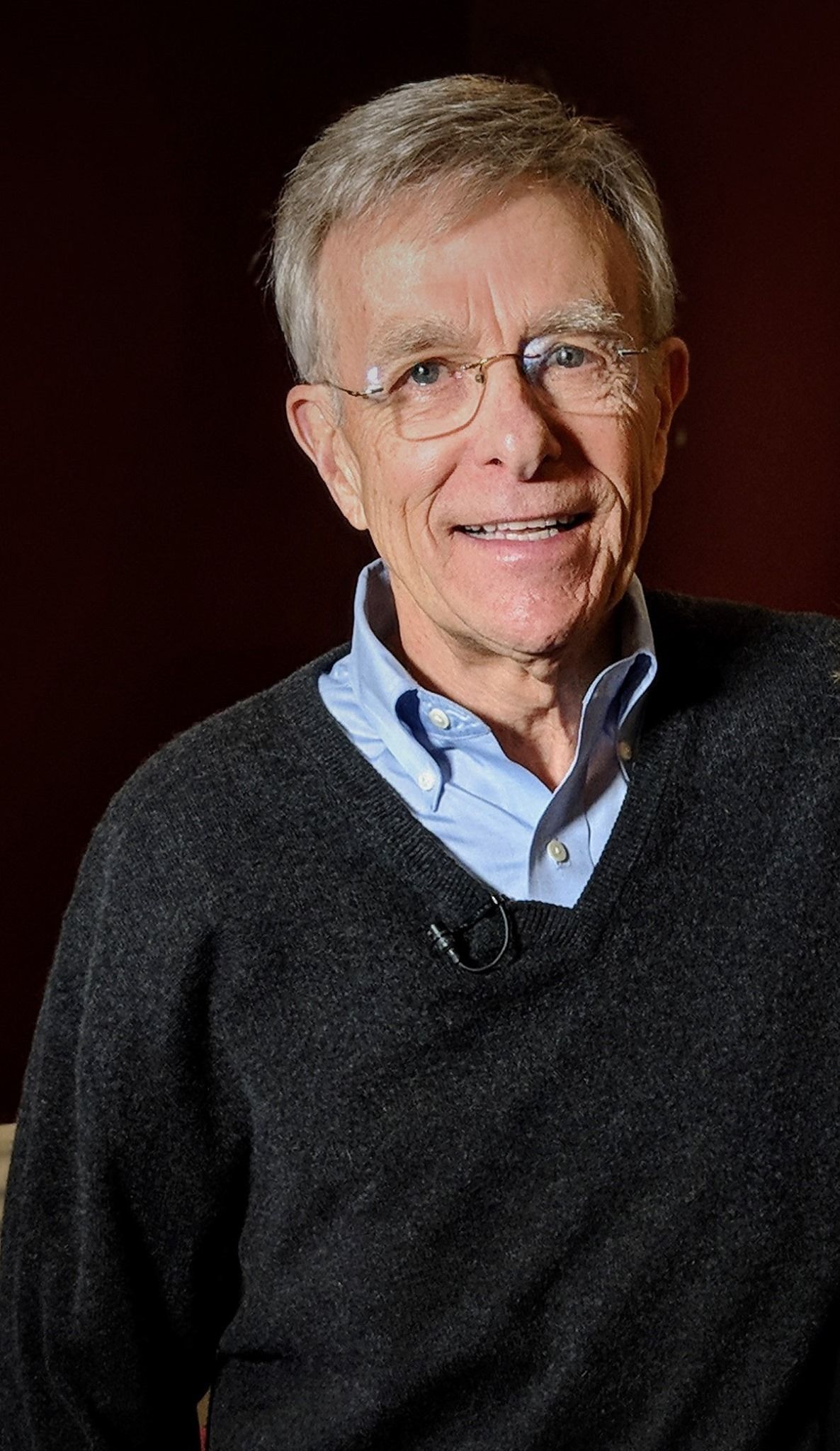 Older man with glasses, wearing blue shirt and sweater, smiling.